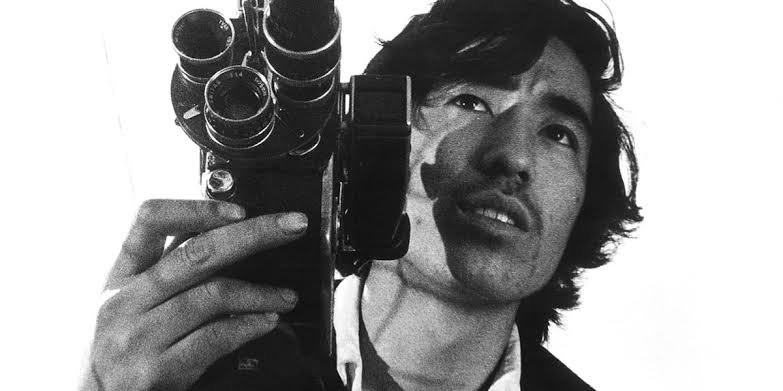 Black and white portrait of a man holding a vintage film camera close to his face against a plain white from background taken from THE MAN WHO LEFT HIS WILL ON FILM.