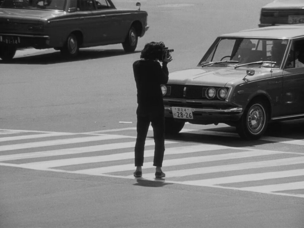 Black and white photograph taken from THE MANO WHO LEF THIS WILL ON FILM, the protagonist standing at a crosswalk holding a camera, with vintage cars on a street in the background.
